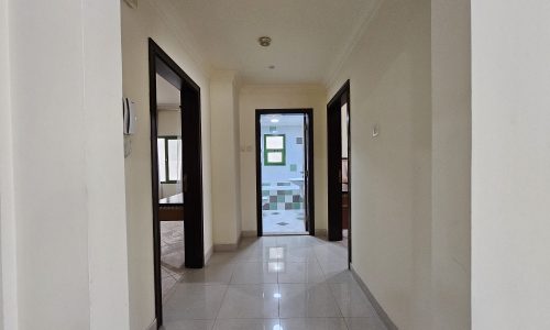 A bright, empty corridor with shiny tiled flooring leads to an open door at the end of a 2BR Apartment for rent in Juffair.