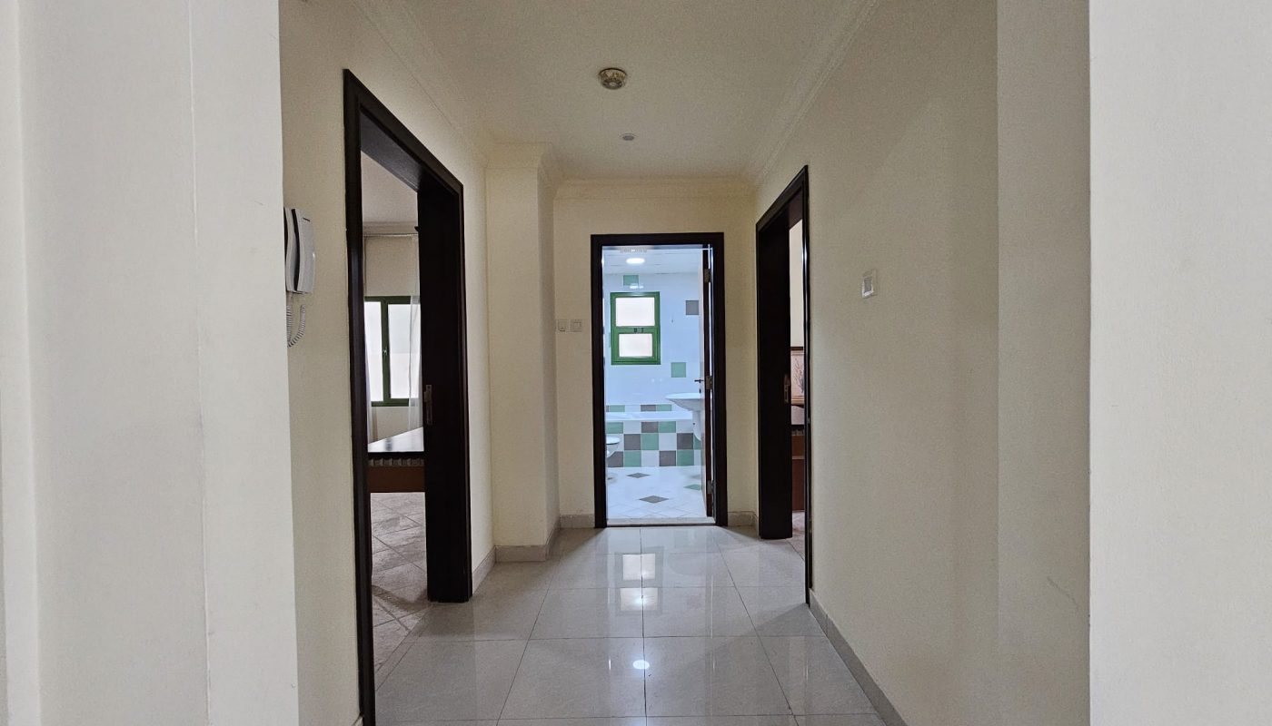 A bright, empty corridor with shiny tiled flooring leads to an open door at the end of a 2BR Apartment for rent in Juffair.