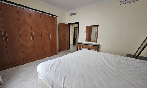 A neatly arranged 2BR apartment in Juffair with a bedroom featuring a large bed, wooden wardrobe, and a dressing table with a mirror, available for rent.