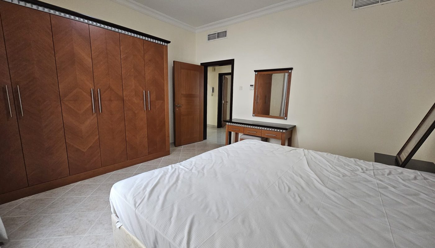 A neatly arranged 2BR apartment in Juffair with a bedroom featuring a large bed, wooden wardrobe, and a dressing table with a mirror, available for rent.