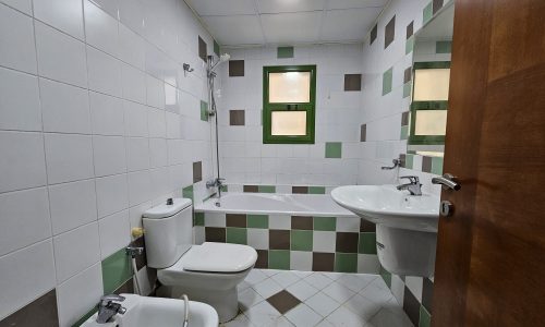 A clean and simple bathroom in a 2BR apartment for rent in Juffair, with white and green tiles, equipped with a toilet, bidet, sink, and bathtub.