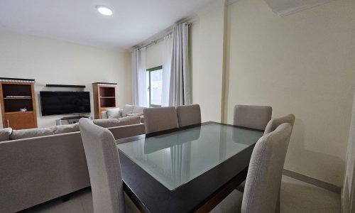 Modern dining room in a Juffair 3 BR apartment for rent, featuring a glass-top table and beige upholstered chairs, adjacent to a living area with a sofa set and entertainment unit.