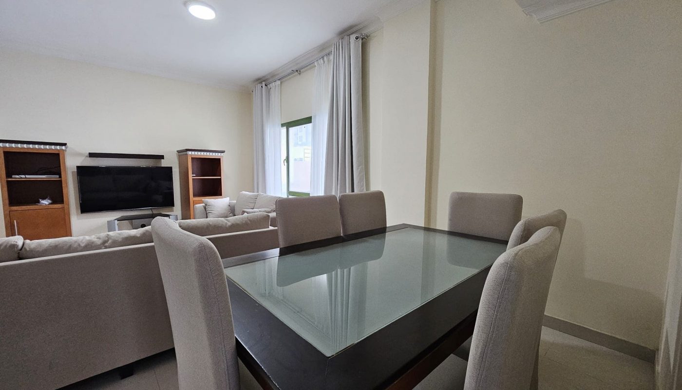 Modern dining room in a Juffair 3 BR apartment for rent, featuring a glass-top table and beige upholstered chairs, adjacent to a living area with a sofa set and entertainment unit.