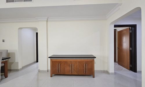 A spacious room corner in a 3 BR apartment for rent in Juffair, with a wooden cabinet and white walls, featuring an archway and a door in the background.