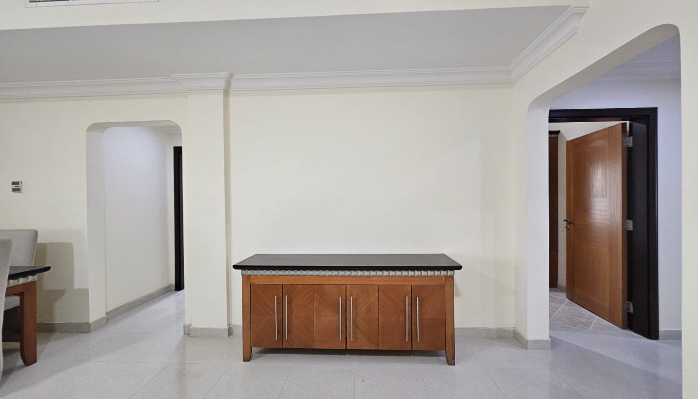 A spacious room corner in a 3 BR apartment for rent in Juffair, with a wooden cabinet and white walls, featuring an archway and a door in the background.
