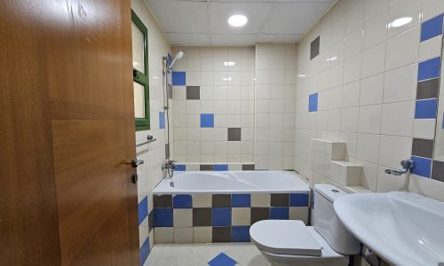 For Rent: A clean, tiled bathroom in a 3 BR Apartment located in Juffair, featuring blue and white tiles, a toilet, two sinks, and a bathtub.