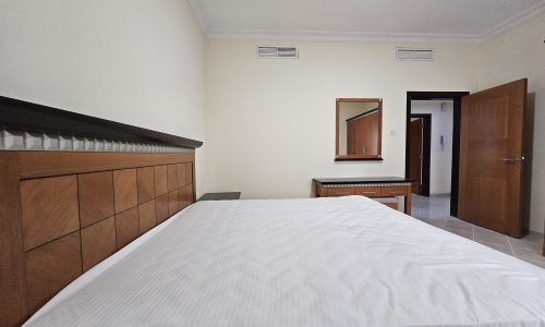 A neatly made bed positioned against a wooden headboard in a tidy bedroom with a mirrored dresser and a door ajar in the background of a 3 BR Apartment for Rent in Juffair.