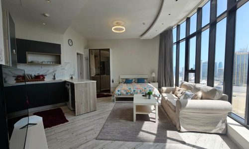 Bright and luxury studio apartment in the Seef Area with large windows and city views, featuring an open-plan living space with a kitchenette, dining area, bed, and sitting area.