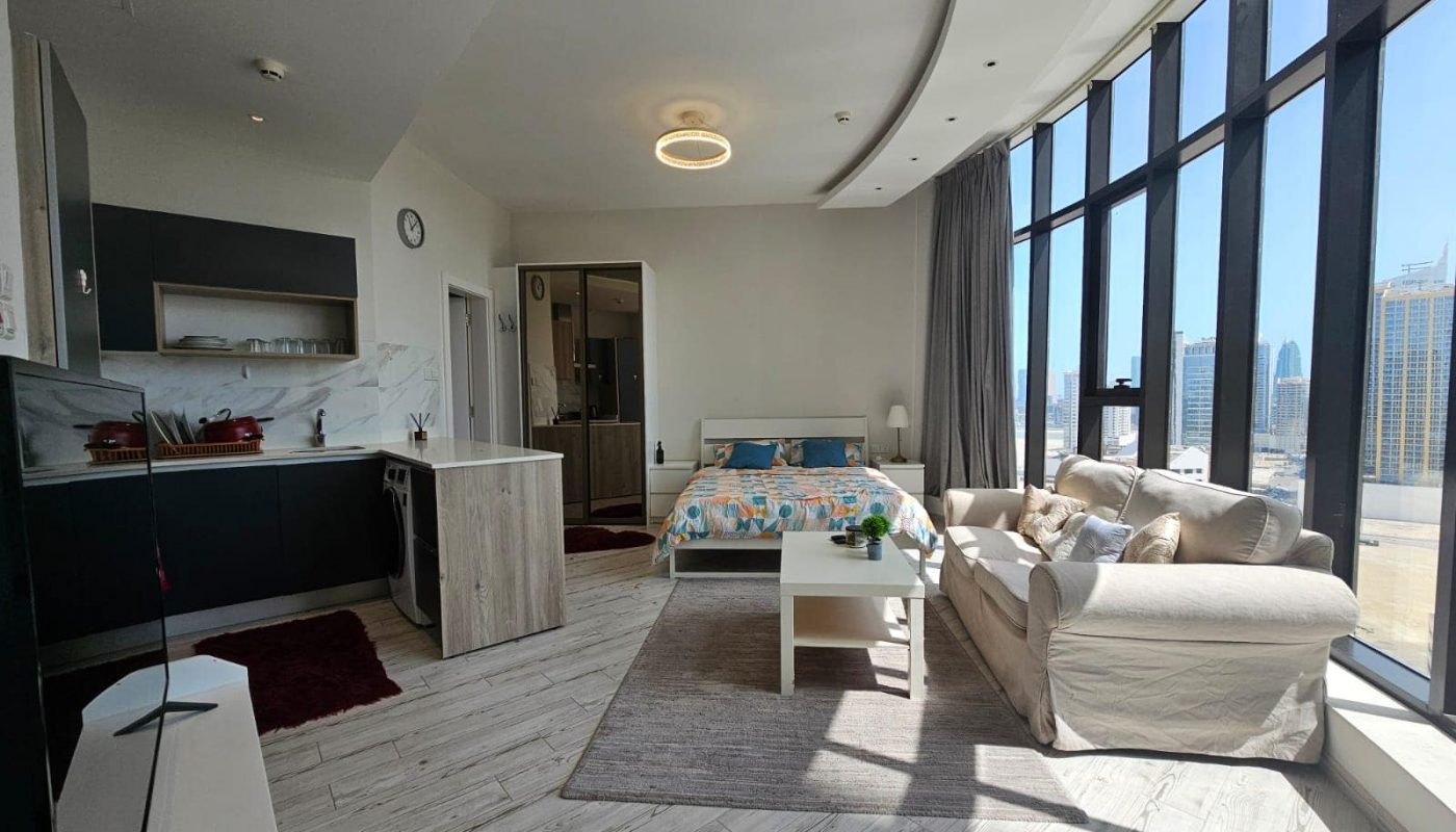 Bright and luxury studio apartment in the Seef Area with large windows and city views, featuring an open-plan living space with a kitchenette, dining area, bed, and sitting area.