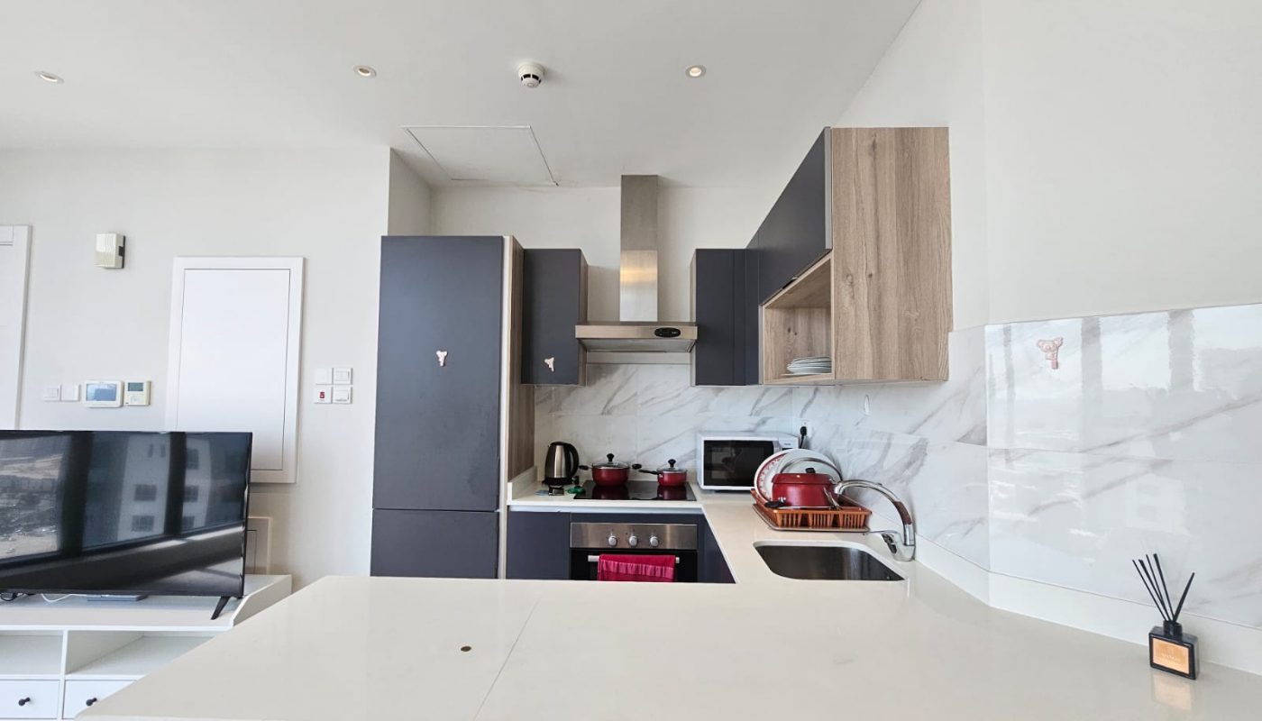 Luxury apartment kitchen interior with marble countertops, wood accents, and gray cabinetry in the Seef area, featuring an island and equipped with appliances for rent.