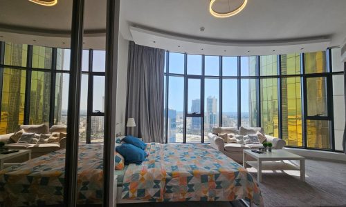 Luxury apartment in the Seef area, with large windows offering a city view, featuring a double bed with colorful bedding and stylish circular lighting fixtures.