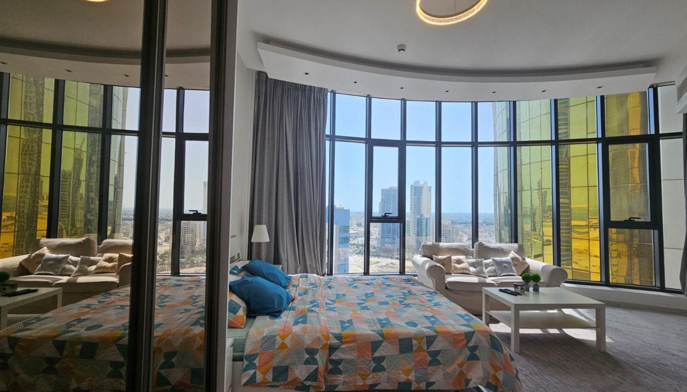Luxury apartment in the Seef area, with large windows offering a city view, featuring a double bed with colorful bedding and stylish circular lighting fixtures.