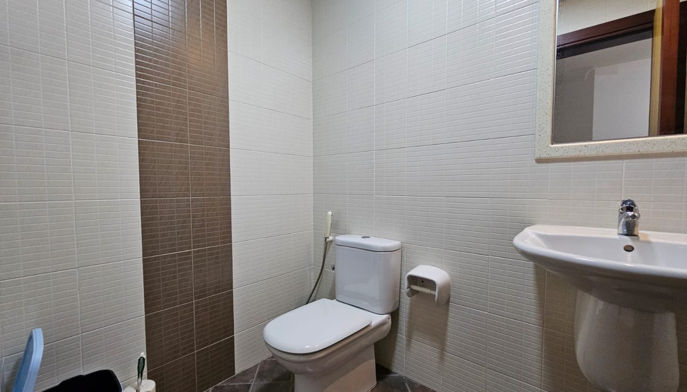 A small, clean bathroom with a toilet, sink, mirror, and tiled walls in a fully-furnished, two-bedroom apartment.
