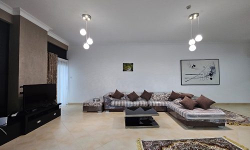 A spacious living room within a fully-furnished two-bedroom apartment in Juffair, featuring low sectional sofas with patterned upholstery, a central black coffee table, a tv set on a console, and