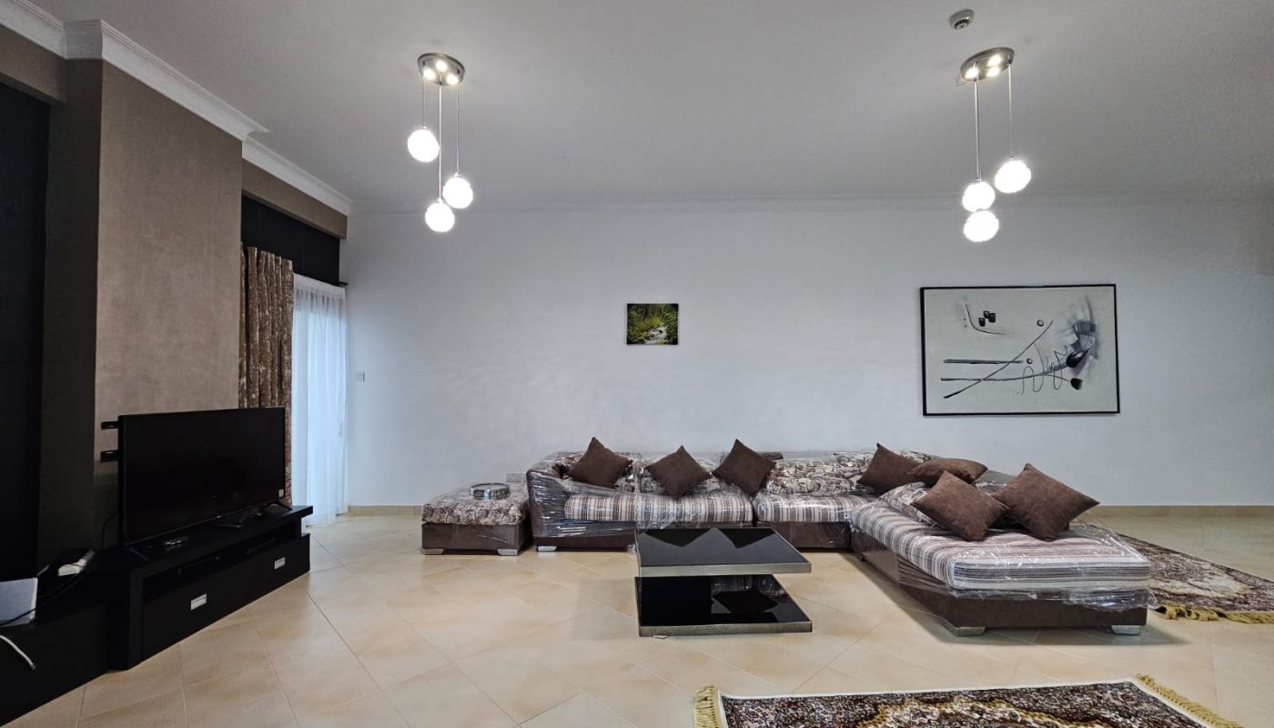 A spacious living room within a fully-furnished two-bedroom apartment in Juffair, featuring low sectional sofas with patterned upholstery, a central black coffee table, a tv set on a console, and