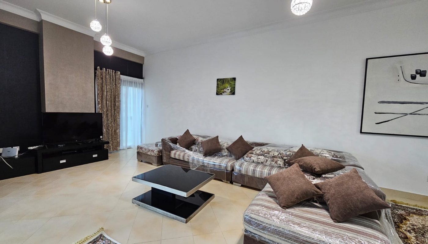 Modern living room in a fully-furnished two-bedroom apartment with sectional sofa, coffee table, tv unit, and decorative lighting in Juffair.