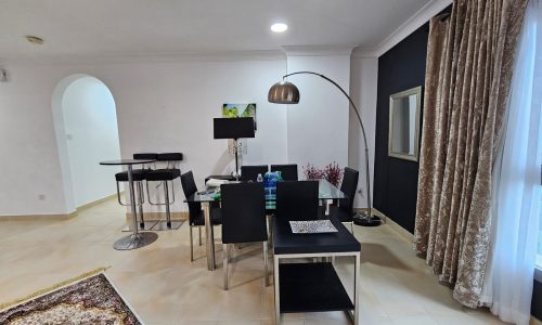 Spacious two-bedroom apartment in Juffair with a modern living room and an open dining area, featuring a patterned area rug, archway, and a mix of natural and artificial lighting.