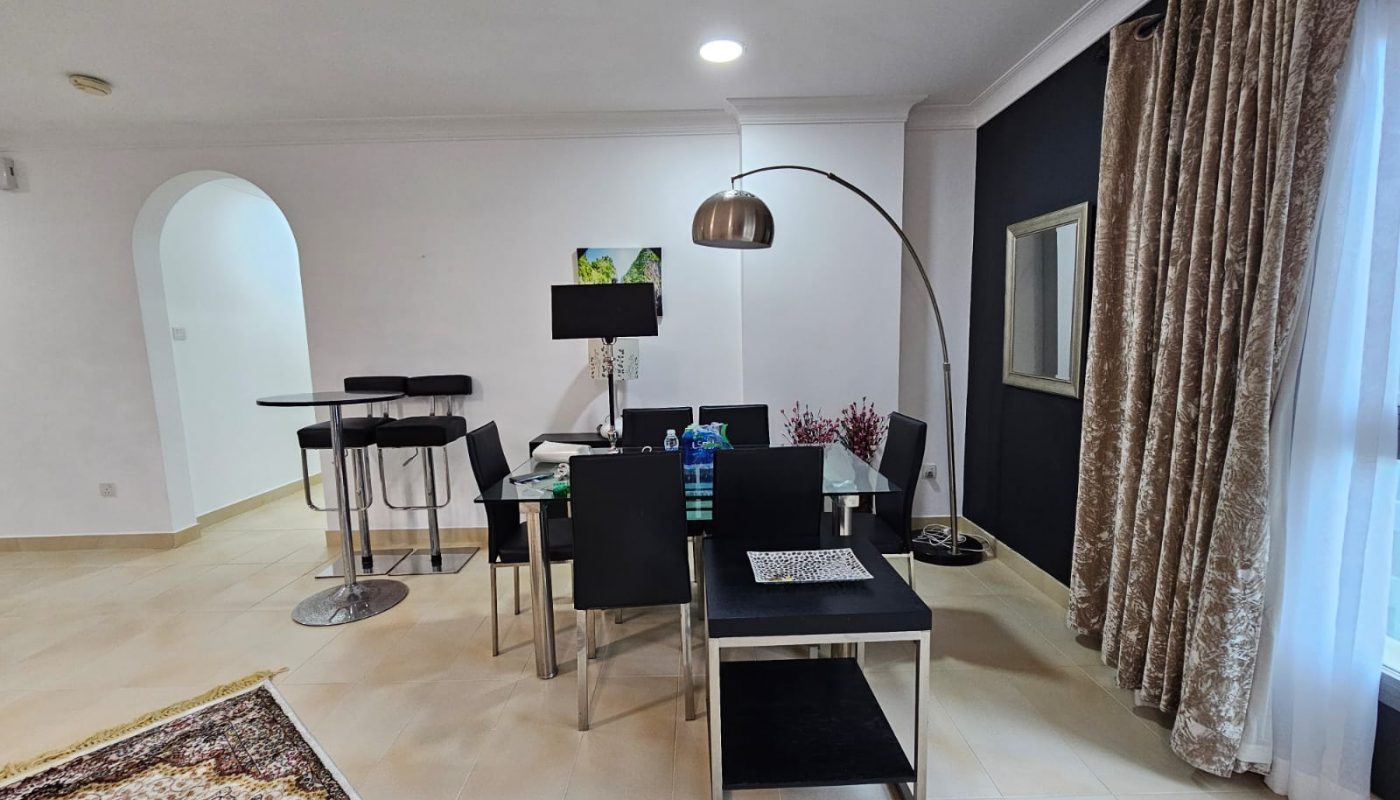 Spacious two-bedroom apartment in Juffair with a modern living room and an open dining area, featuring a patterned area rug, archway, and a mix of natural and artificial lighting.