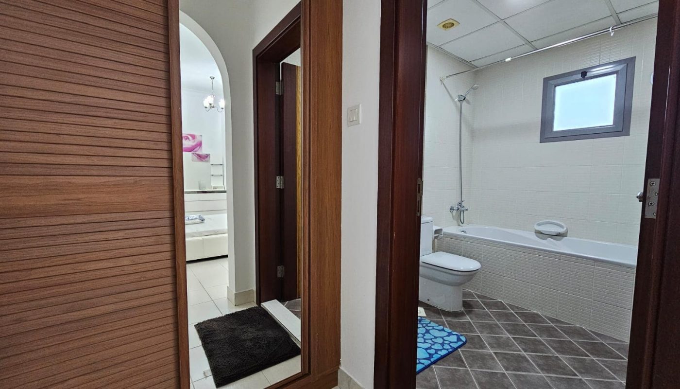 A view into a furnished, modern bathroom in a two-bedroom apartment in Juffair through an open door showing a toilet, a sink, and a mirror.
