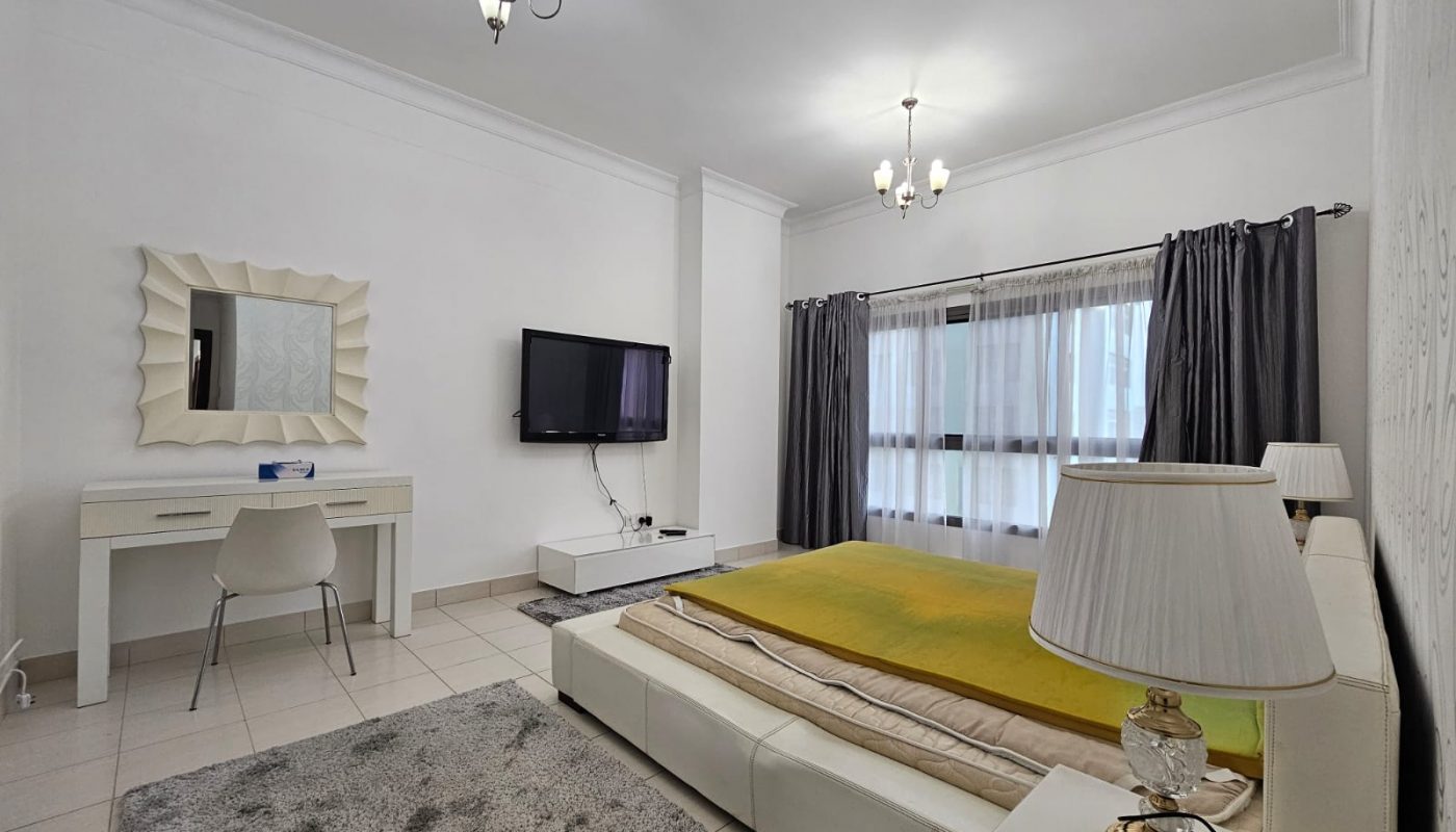 Bright, modern TWO BEDROOM apartment with a minimalist design in Juffair, featuring large beds with yellow covers, televisions, desks, and elegant lighting.