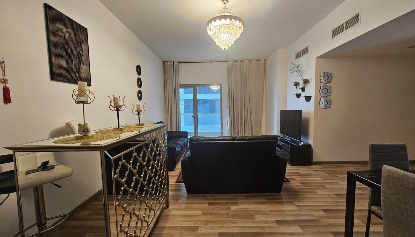 A neatly arranged living room in an elegant apartment for rent in the Juffair area, with a black sofa set, a glass coffee table, and decorative wall art, leading to a balcony with curtains