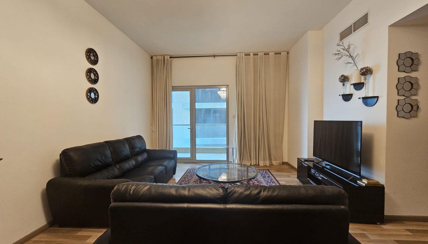 An elegant apartment for rent showcasing a modern living room with a black sofa set, a glass coffee table, a television, and decorative wall plates, all complemented by a view to a balcony in J