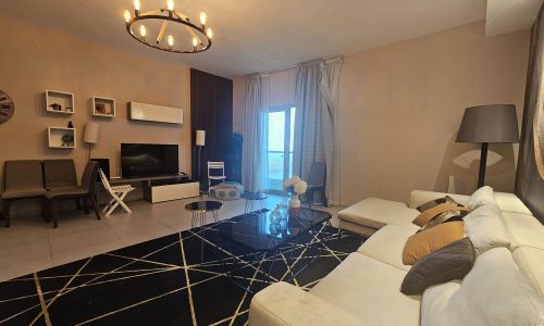 Modern living room in a Juffair flat for rent with neutral colors, featuring a sofa, armchairs, a television, and decorative lighting.