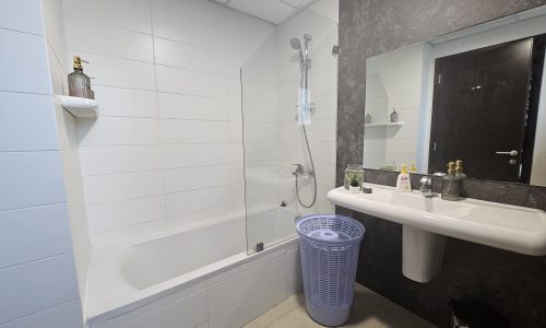 A modern bathroom interior in a Juffair flat for rent, featuring a bathtub and shower combination, white wall tiles, a sink, and various toiletries.