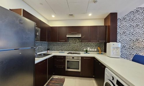 A modern kitchen in an apartment for rent in Juffair with stainless steel appliances, dark wood cabinets, and a decorative tiled backsplash.