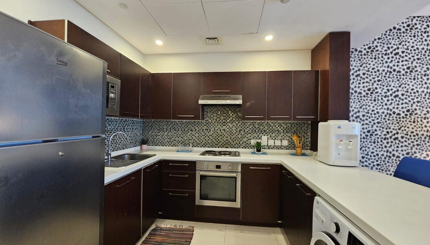 A modern kitchen in an apartment for rent in Juffair with stainless steel appliances, dark wood cabinets, and a decorative tiled backsplash.