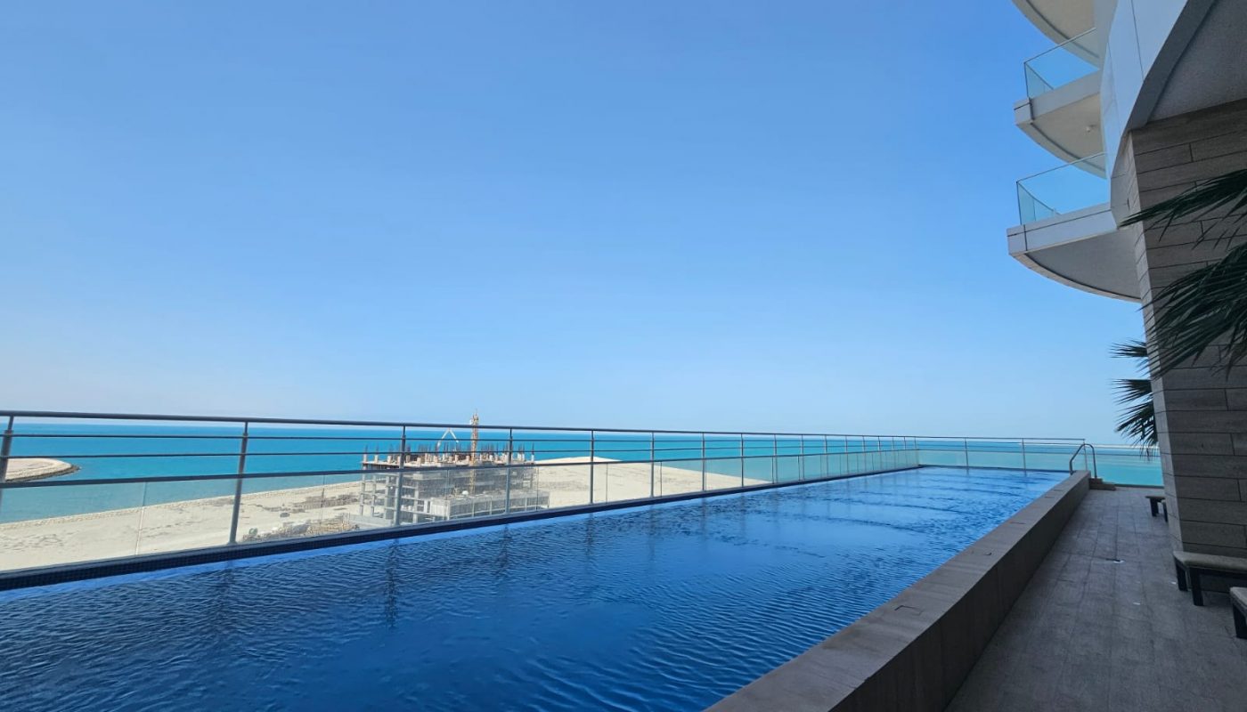 Infinity pool on a high terrace overlooks the beach and ocean, with clear blue sky and part of a modern building visible on the right.
