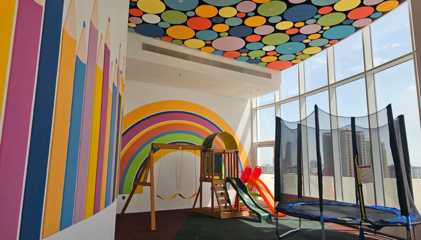 Indoor playroom with colorful wall and ceiling designs, featuring a swing set, slide, trampoline, and large windows letting in natural light.