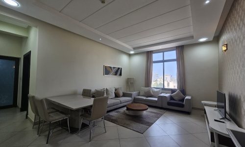 Modern living room with dining area in a 2BR Furnished Flat, featuring a neutral color palette, couch, dining table, and TV, illuminated by natural and artificial light.