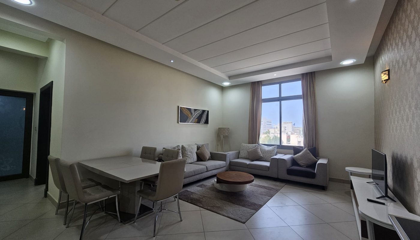Modern living room with dining area in a 2BR Furnished Flat, featuring a neutral color palette, couch, dining table, and TV, illuminated by natural and artificial light.