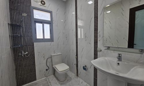 A modern bathroom interior in a 2BR Furnished Flat in Adliya, featuring white and gray marble tiles, a glass shower enclosure, a white toilet, and a pedestal sink.