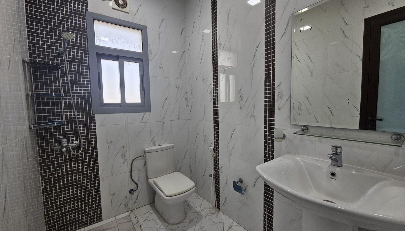 A modern bathroom interior in a 2BR Furnished Flat in Adliya, featuring white and gray marble tiles, a glass shower enclosure, a white toilet, and a pedestal sink.