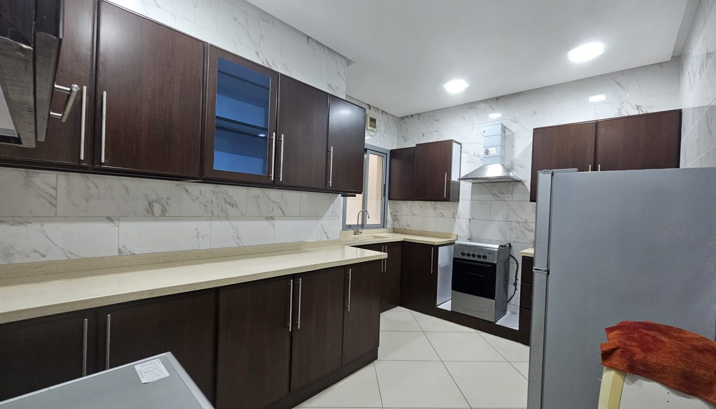 A modern kitchen in a 2BR furnished flat with wooden cabinets, marble countertops, and stainless steel appliances, located in Adliya.