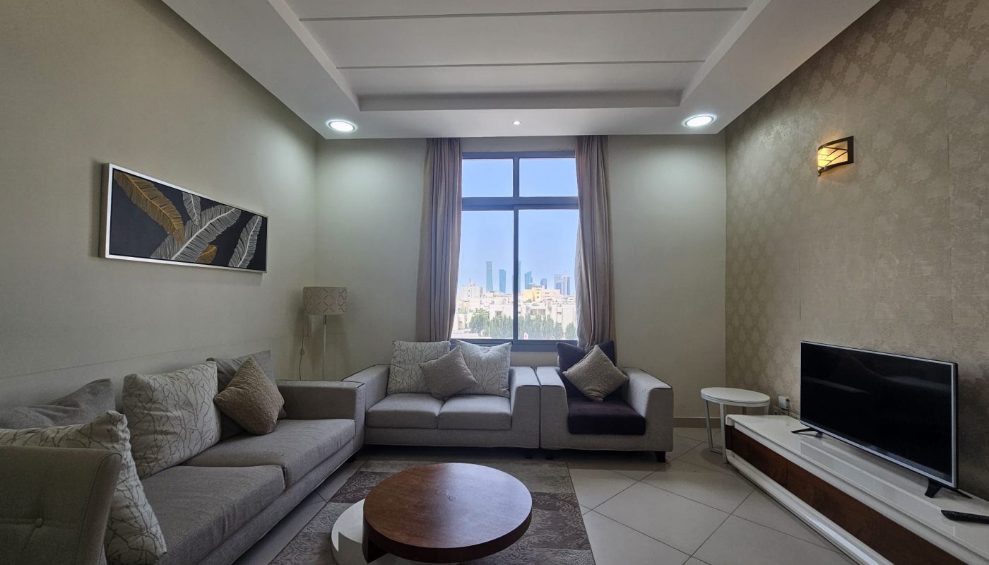 Modern living room in a 2BR Adliya furnished flat with a neutral color scheme, featuring a sofa, armchair, and television, with a cityscape view through the window.