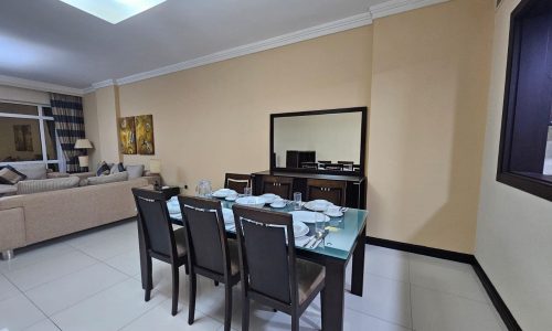 A modern 3BR apartment featuring a dining area with a set table, adjacent to a living space with a large TV and sofa.