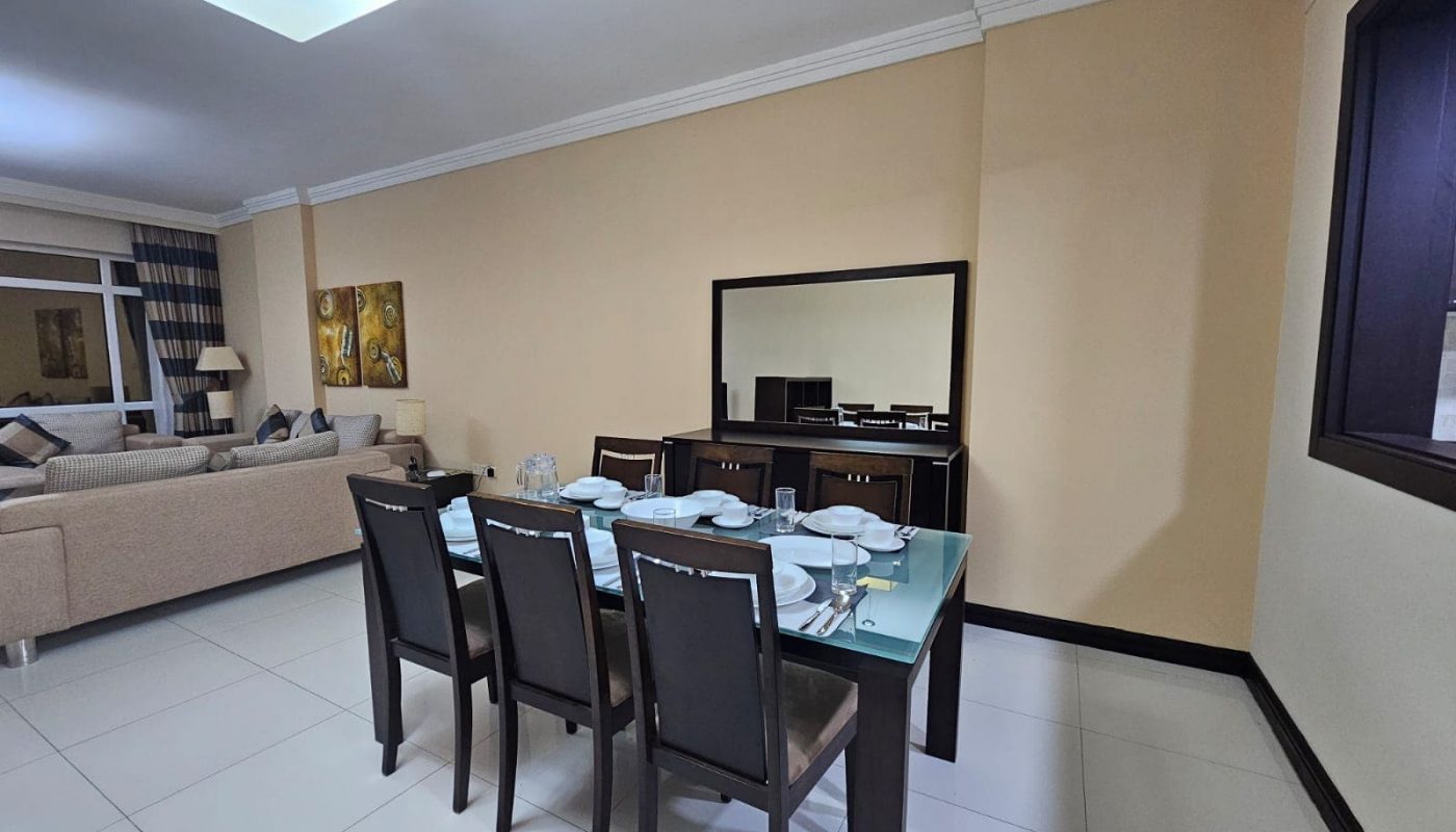 A modern 3BR apartment featuring a dining area with a set table, adjacent to a living space with a large TV and sofa.