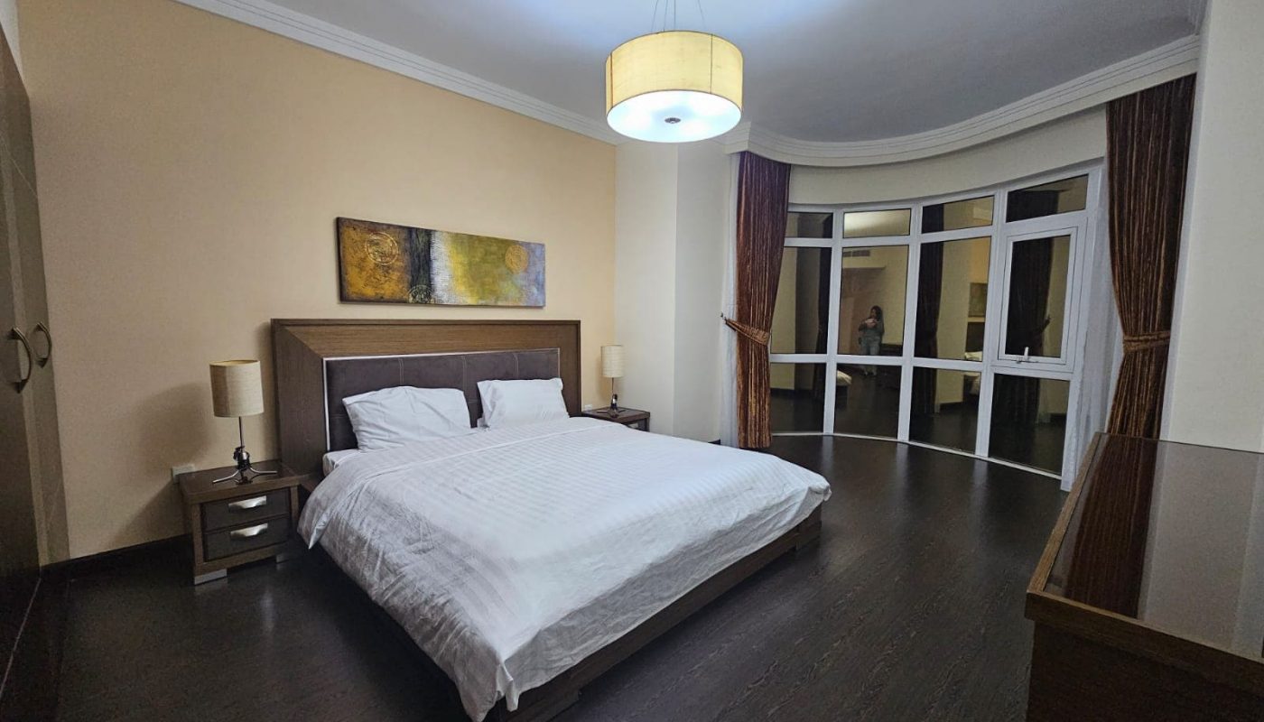 A well-lit, modern 3BR apartment bedroom with a double bed, nightstands, and a person standing by the French doors.