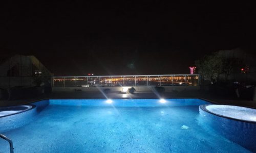 An illuminated swimming pool at night with modern 3BR apartment buildings in the background.