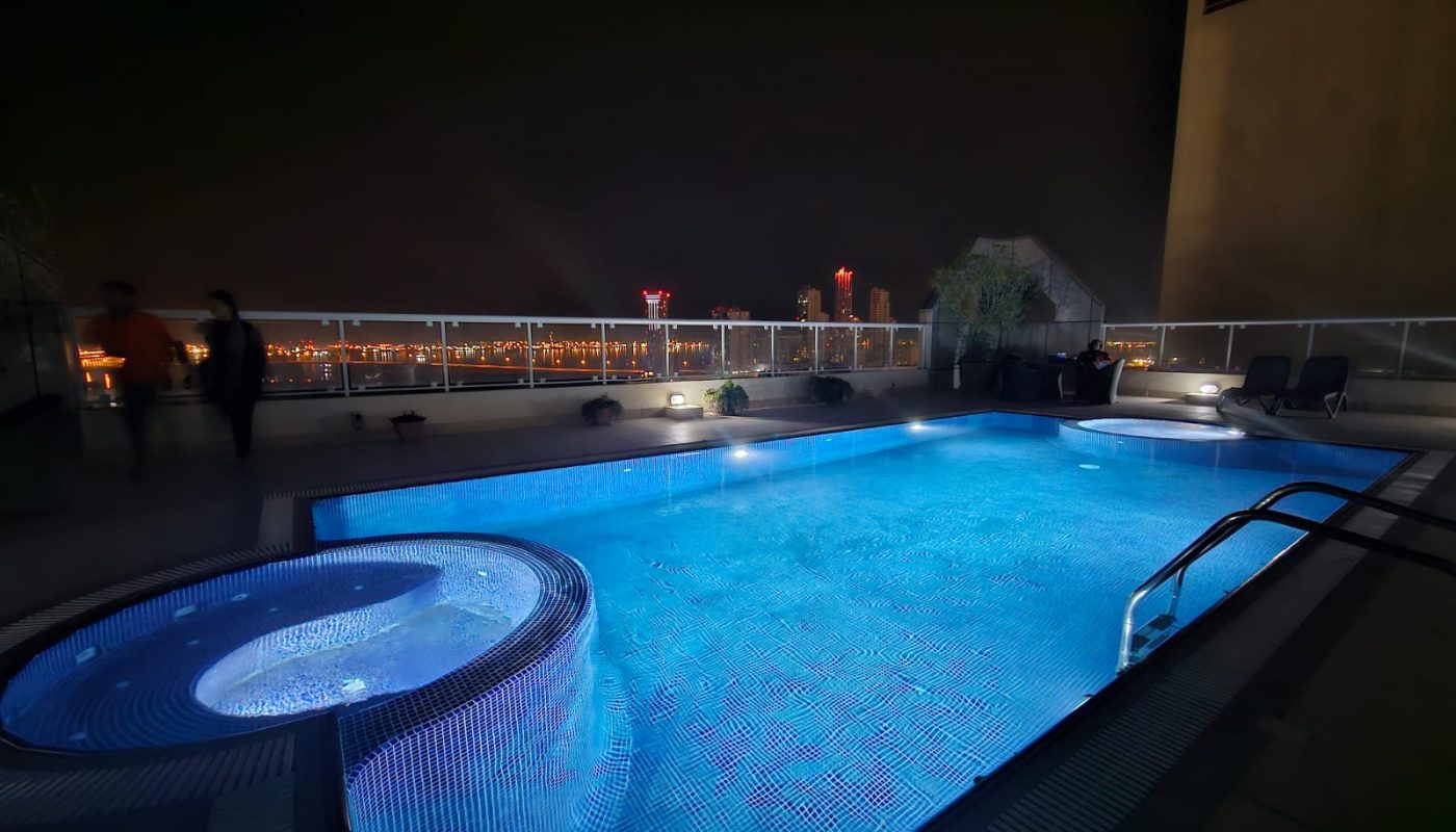 Rooftop pool with blue lighting at nighttime, city lights in the background of a modern 3BR apartment.