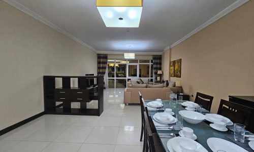 Modern 3BR apartment with a dining area featuring a set table, leading into a living space with beige decor and a chandelier.