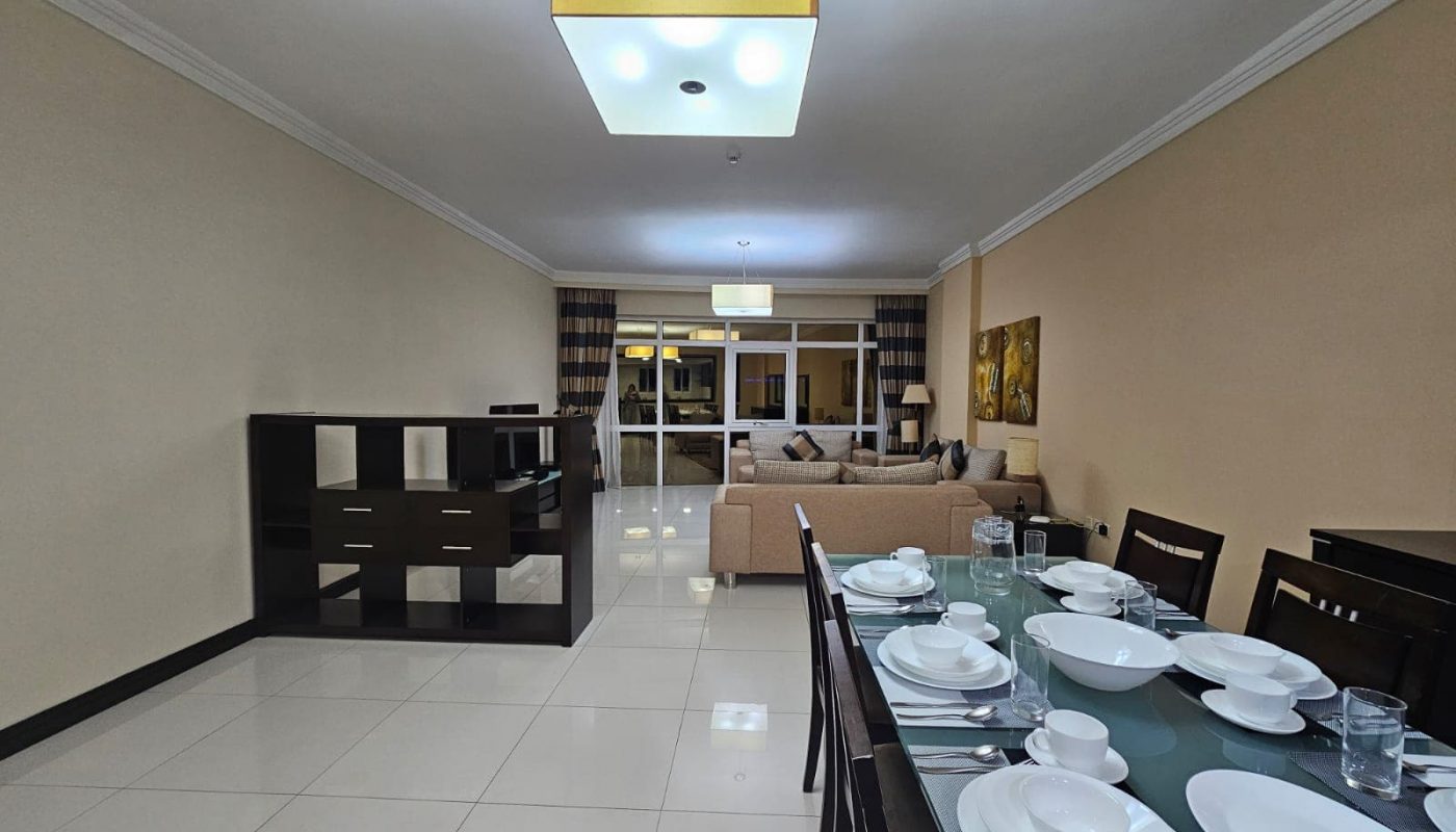 Modern 3BR apartment with a dining area featuring a set table, leading into a living space with beige decor and a chandelier.