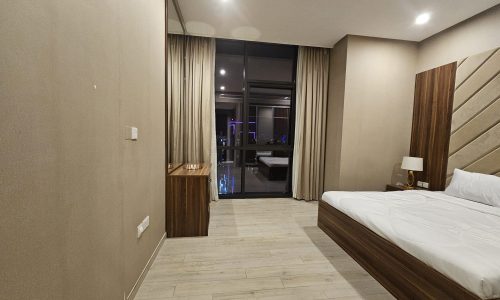 Modern one-bedroom apartment featuring a large bed, wooden headboard, beige walls, curtains, and an adjoining room with ambient lighting visible through the glass door.