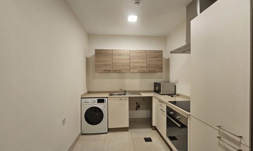 A modern, fully-furnished kitchen with white cabinets, a washing machine, and a wooden countertop under recessed lighting.