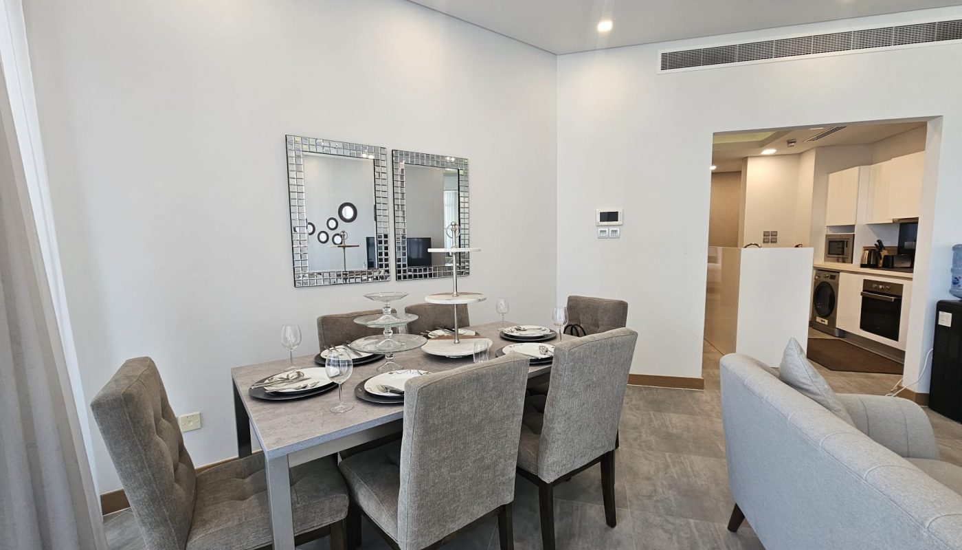 Modern dining area with a table set for four, cushioned chairs, and mirrors on the wall. The adjoining kitchen is visible in the background.