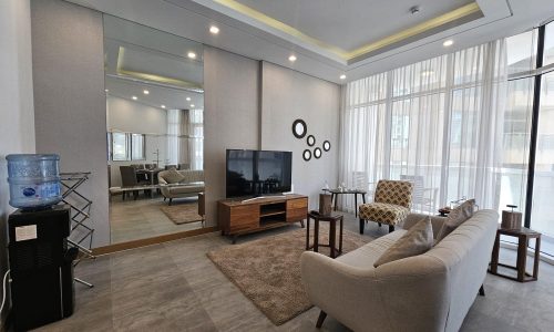 Modern living room in a Juffair flat rental with grey sofa, patterned armchair, and TV on wooden stand, with ambient ceiling lighting and large windows.