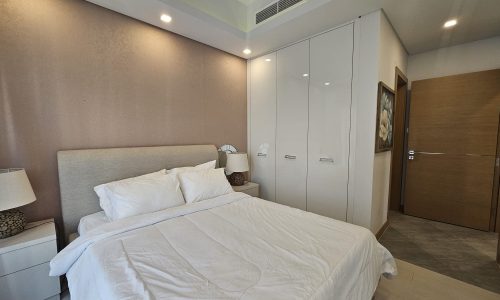 Modern flat bedroom with a large bed, beige headboard, white bedding, two bedside lamps, a large wardrobe, and a wooden door.